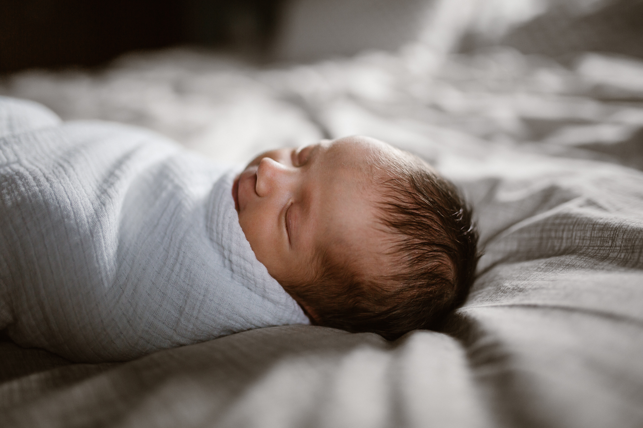 Newborn swaddled in a white blanket, sleeping on the bed in window light.