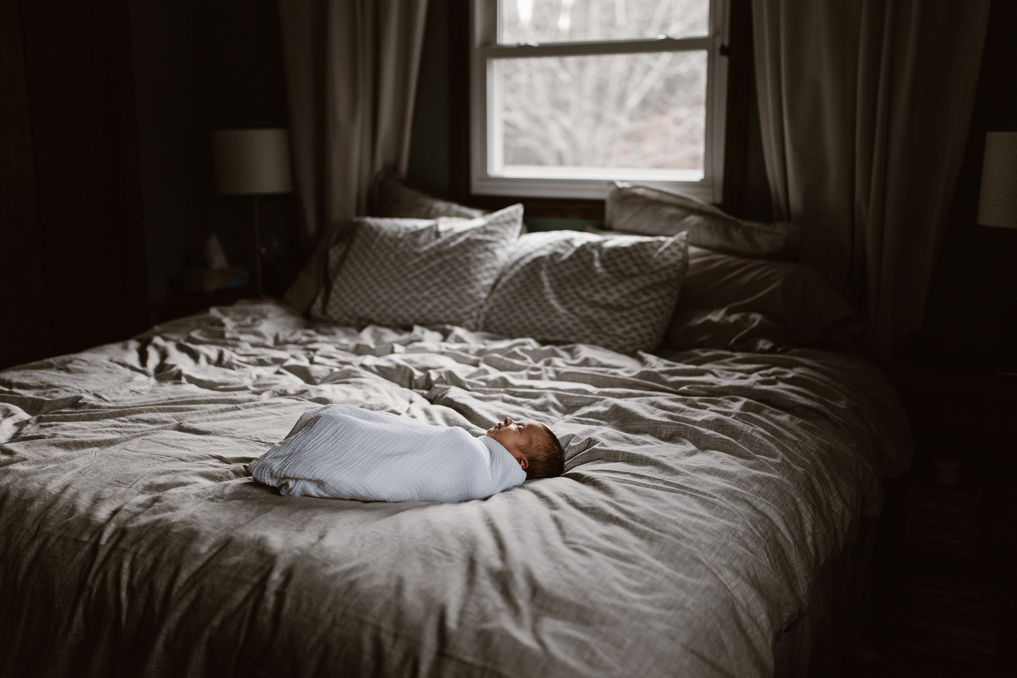 Newborn swaddled on a bed in moody window light.