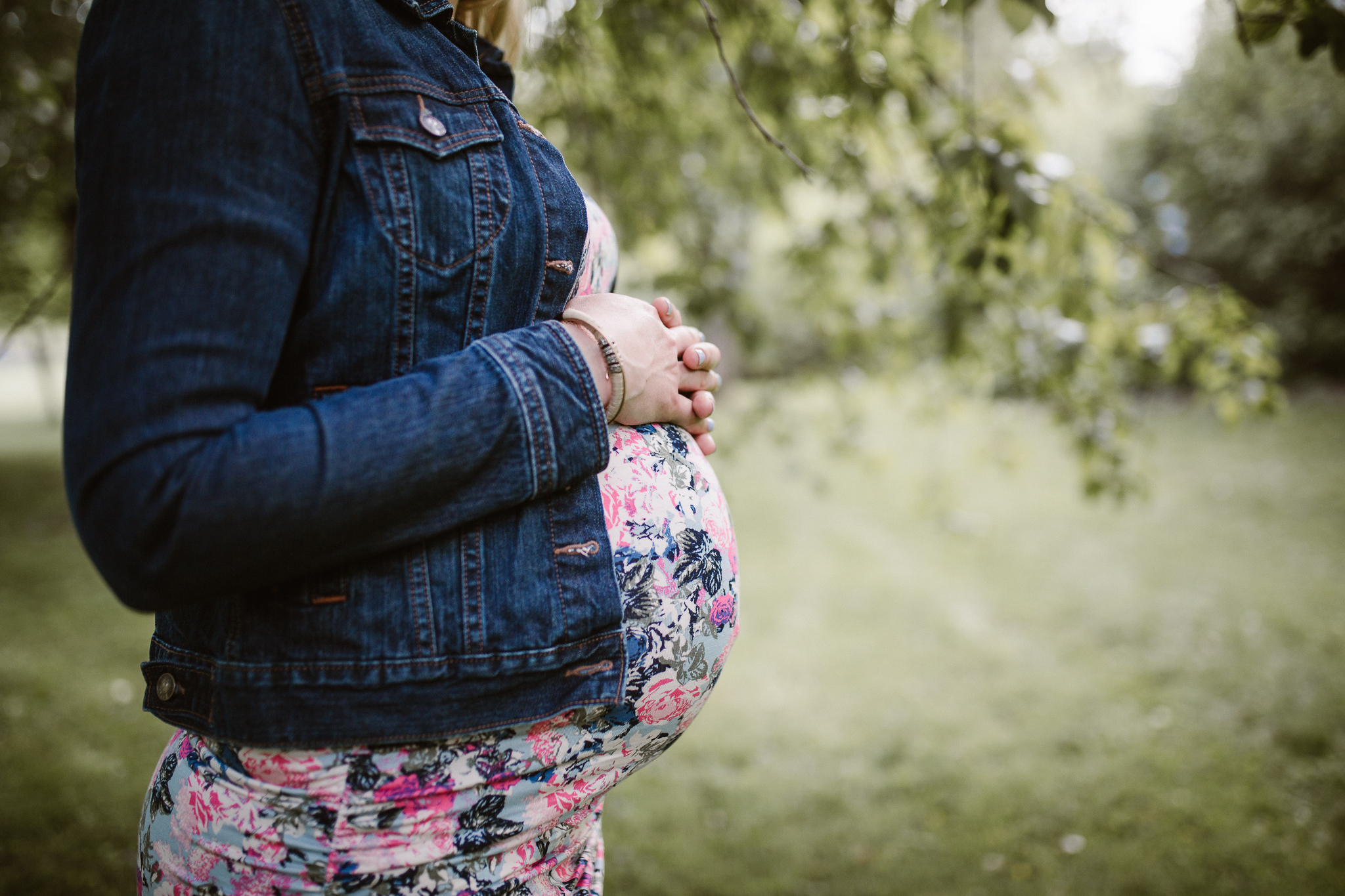 Pregnant mother in denim holding the top of her belly amongst the spring tree leaves.