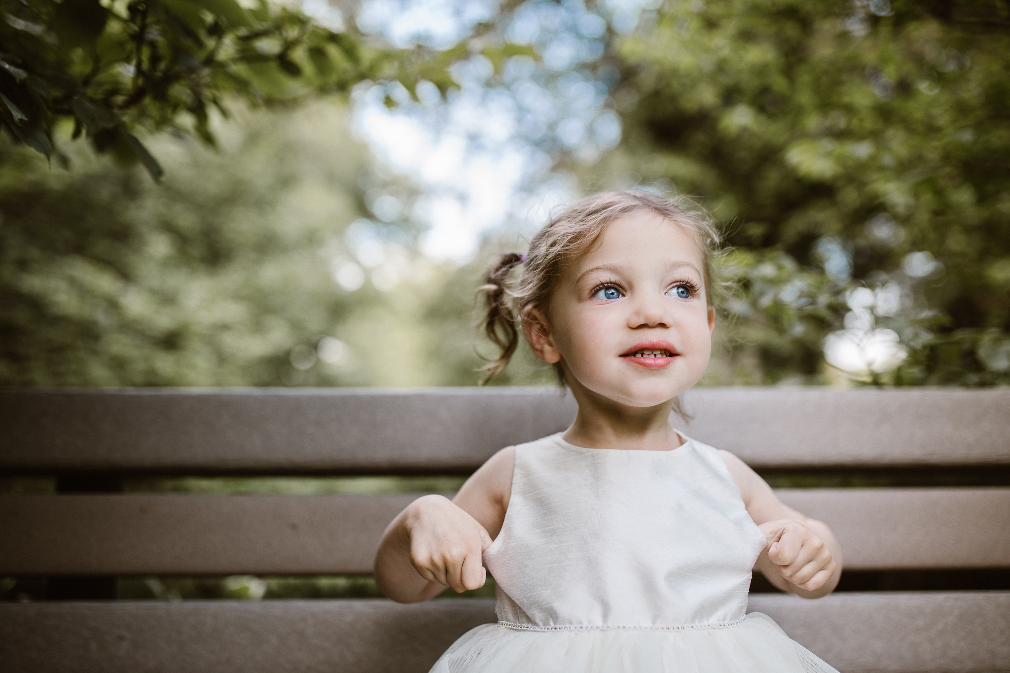 Toddler girl sitting outside on a wooden bench pulling at her white dress.