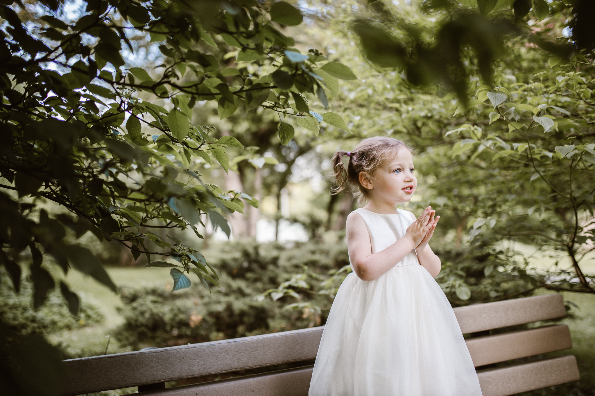 Toddler girl standing on a wooden bench in the foliage.