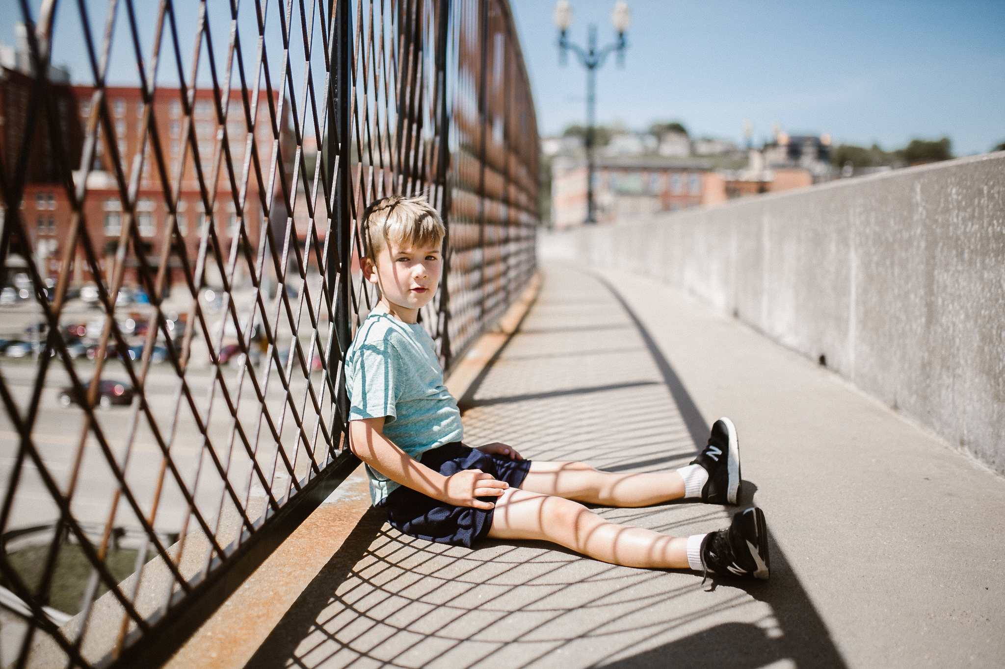 Boy sitting down in harsh light against a bridge fence which casts shadows.