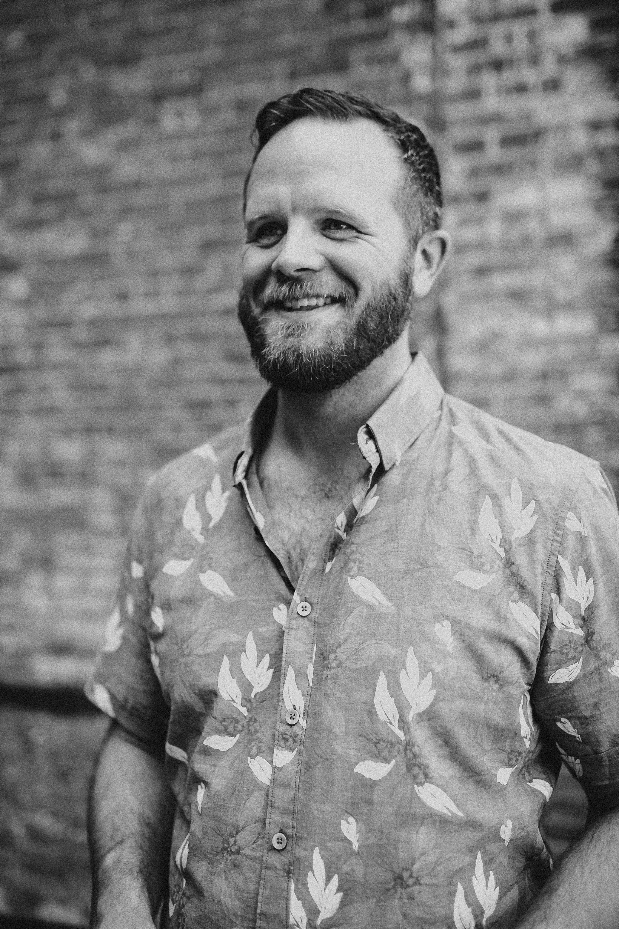 Handsome man smiling in front of a brick building wearing a floral shirt.