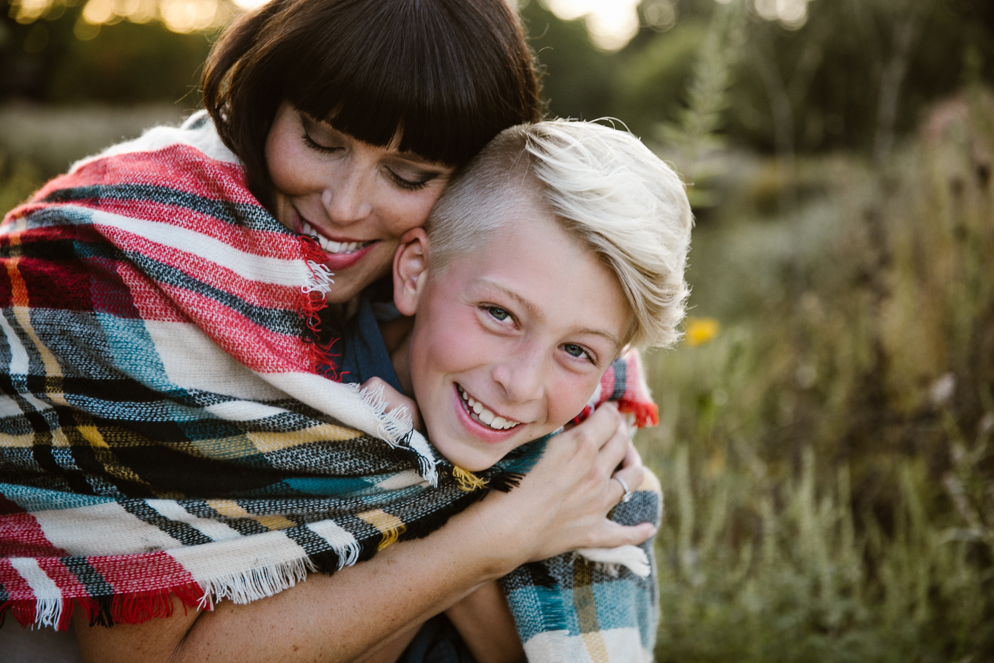 Mother wrapping a plaid blanket around her tween son, hugging him lovingly at sunset.