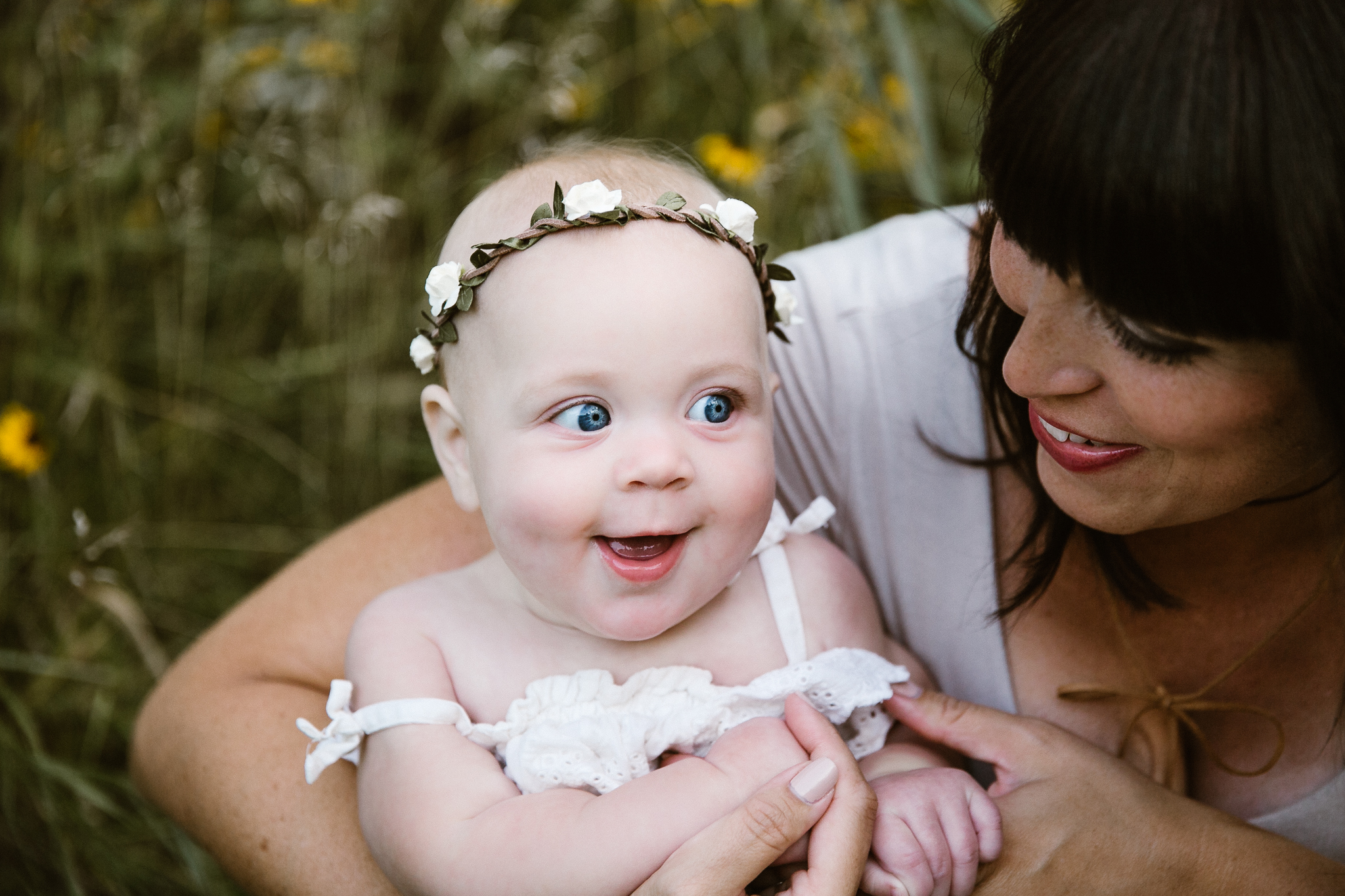 Baby girl with a cute smile and flower crown sitting on her mother's lap in wildflowers.