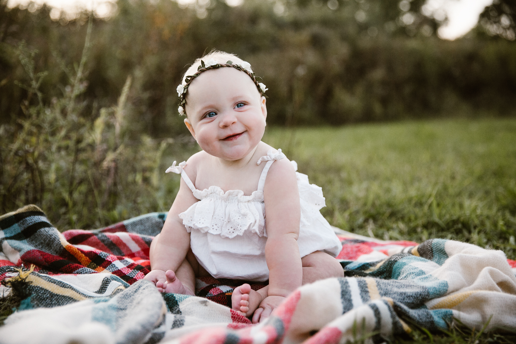 Portrait of a baby girl sitting up on a plaid blanket at sunset.