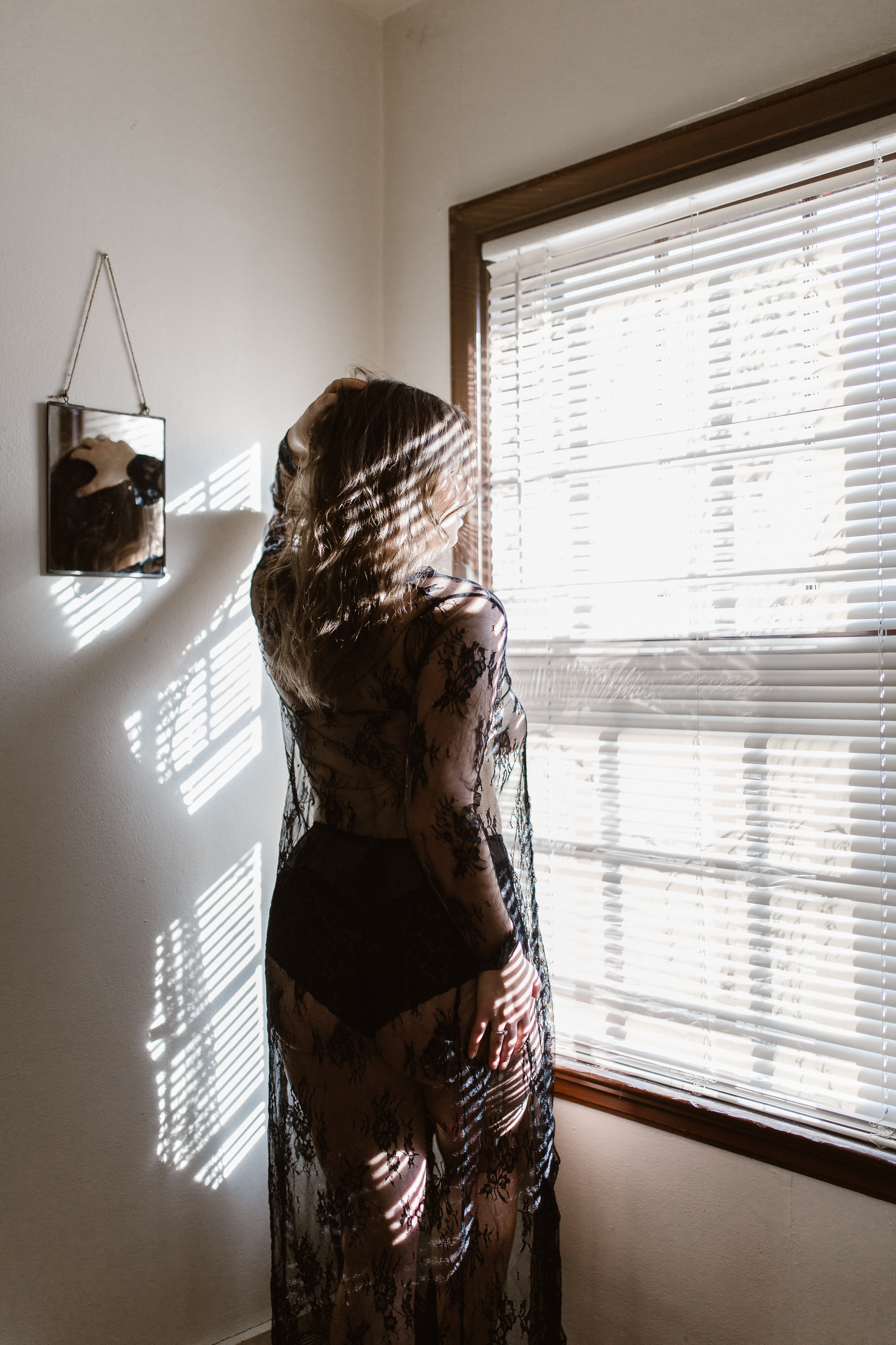 Intimate boudoir shot of woman in a black, lace robe standing in front of window blind shadows.